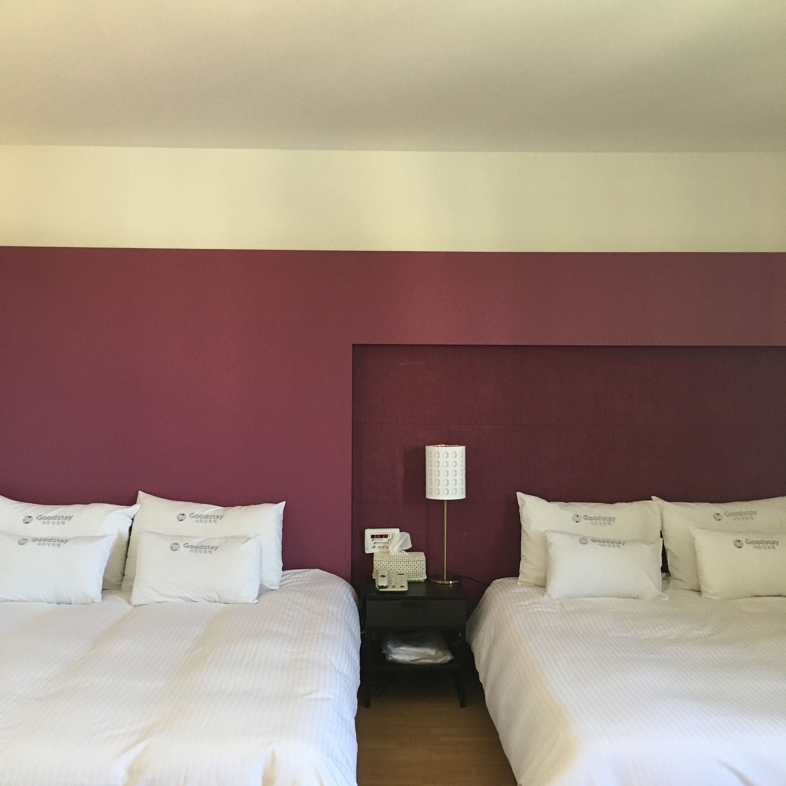 A hotel room with two beds, each with white bedding and pillows labeled "Goodstay." There is a nightstand with a lamp and a tissue box between the beds.