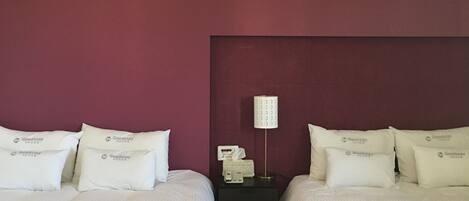 A hotel room with two beds, each with white bedding and pillows labeled "Goodstay." There is a nightstand with a lamp and a tissue box between the beds.