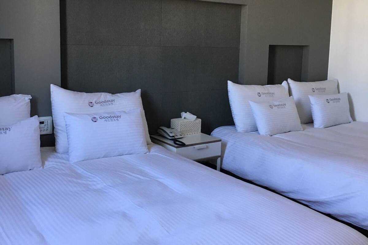 Une chambre d’hôtel avec deux lits, chacun équipé de draps et d’oreillers blancs portant la mention « Goodstay ». On y trouve une table de chevet avec un téléphone et une boîte de mouchoirs.
