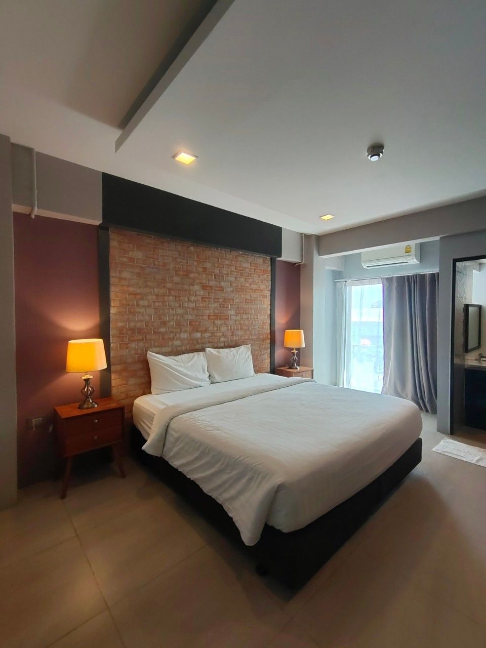 A hotel room with a large bed, two bedside tables with lamps, a brick accent wall, an air conditioning unit, and a view of the outdoors through a window.