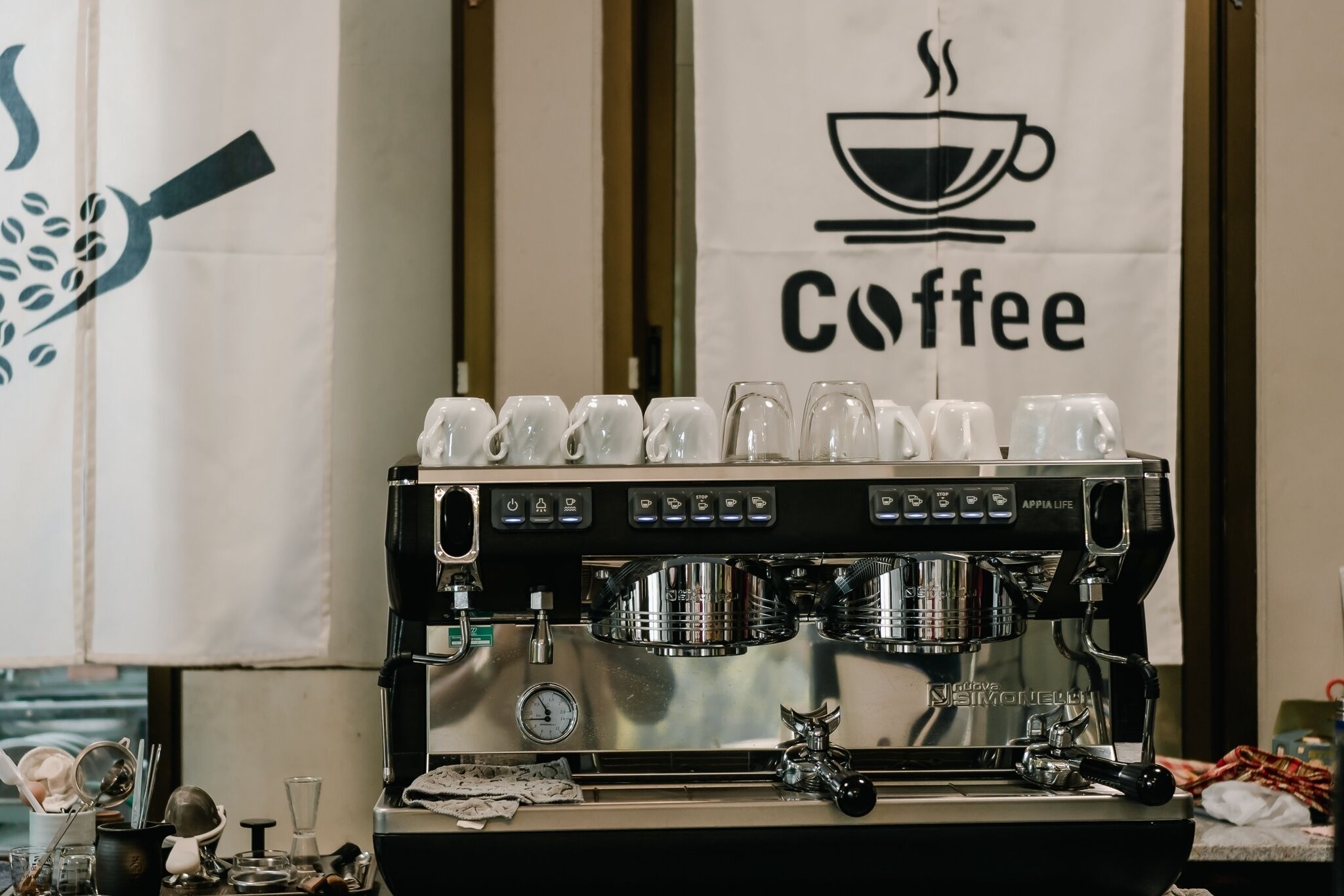 A coffee machine with multiple cups stacked on top, a coffee banner, and a window in the background.