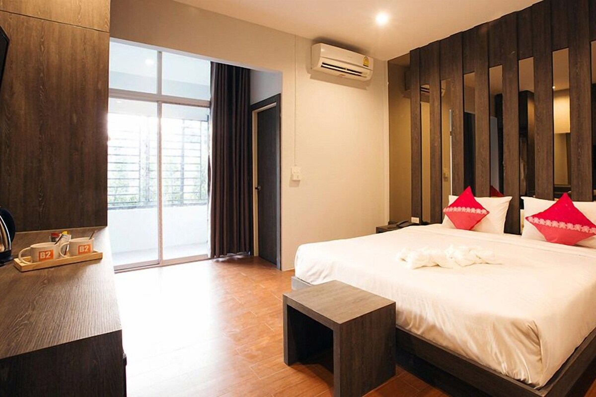 A hotel room with a large bed, a desk with a kettle, a small table, and a sliding door to a balcony.