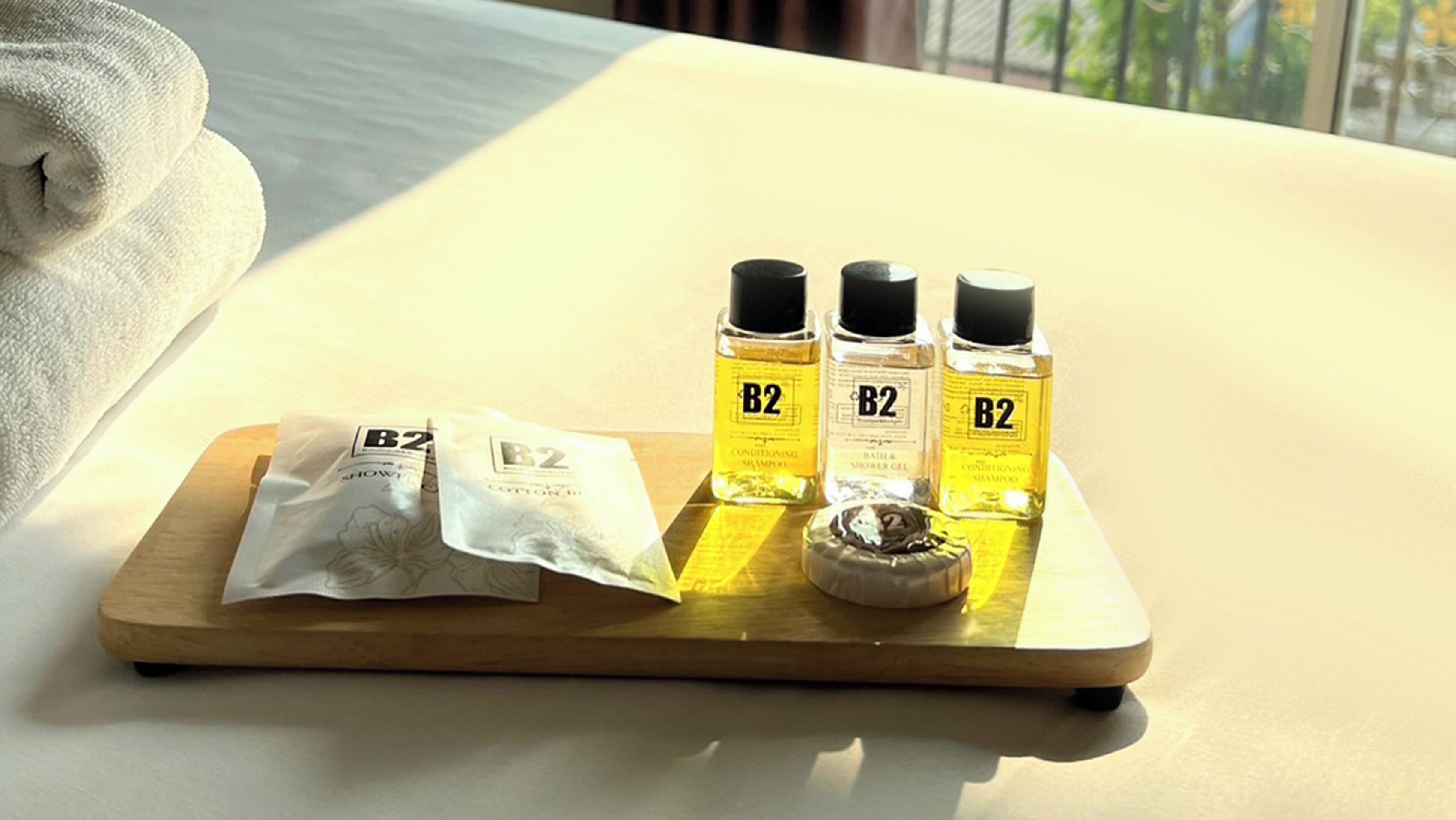 A wooden tray with three bottles of B2 conditioning shampoo, a packet of B2 show shower gel, and a packet of B2 conditioning shampoo on a table by a window.