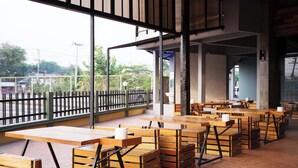 An outdoor seating area with wooden tables and benches, a brick wall, and a view of greenery and buildings in the background. - B2 Buriram Boutique & Budget Hotel (Buriram)