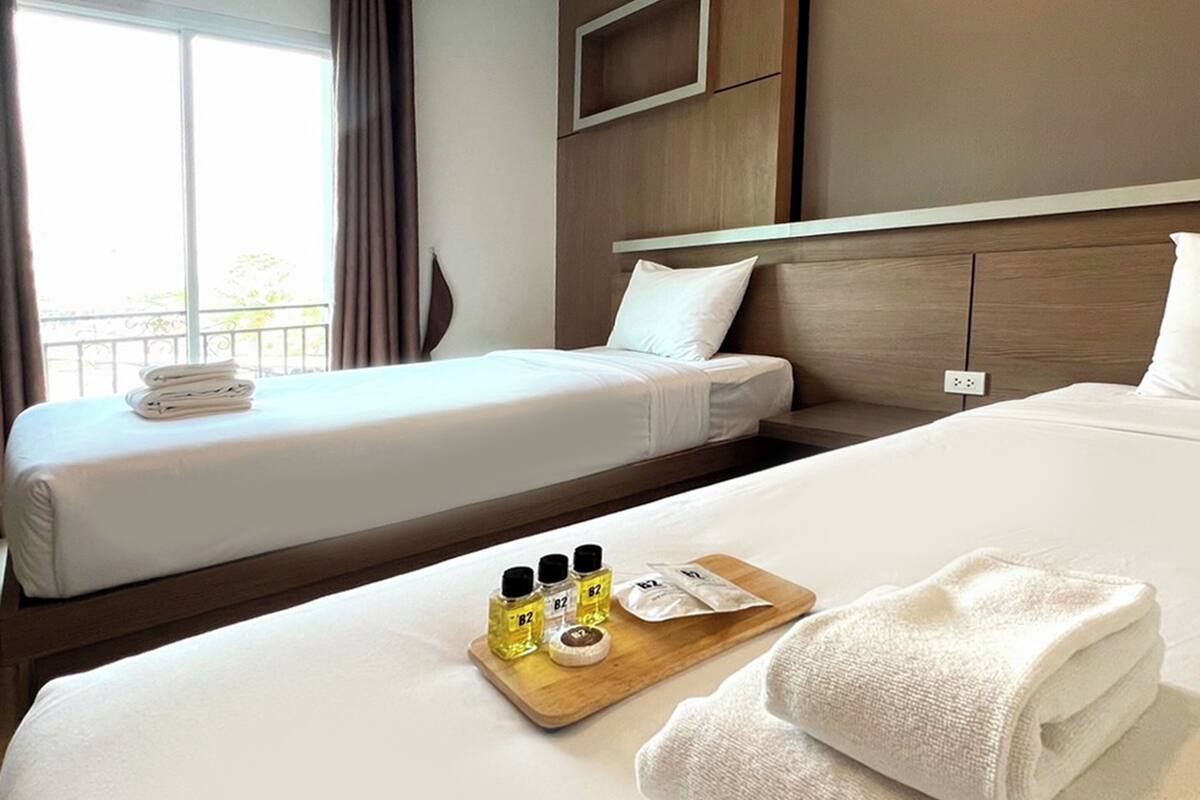 A hotel room with two beds, a wooden headboard, a bedside table with toiletries, and a window with curtains.