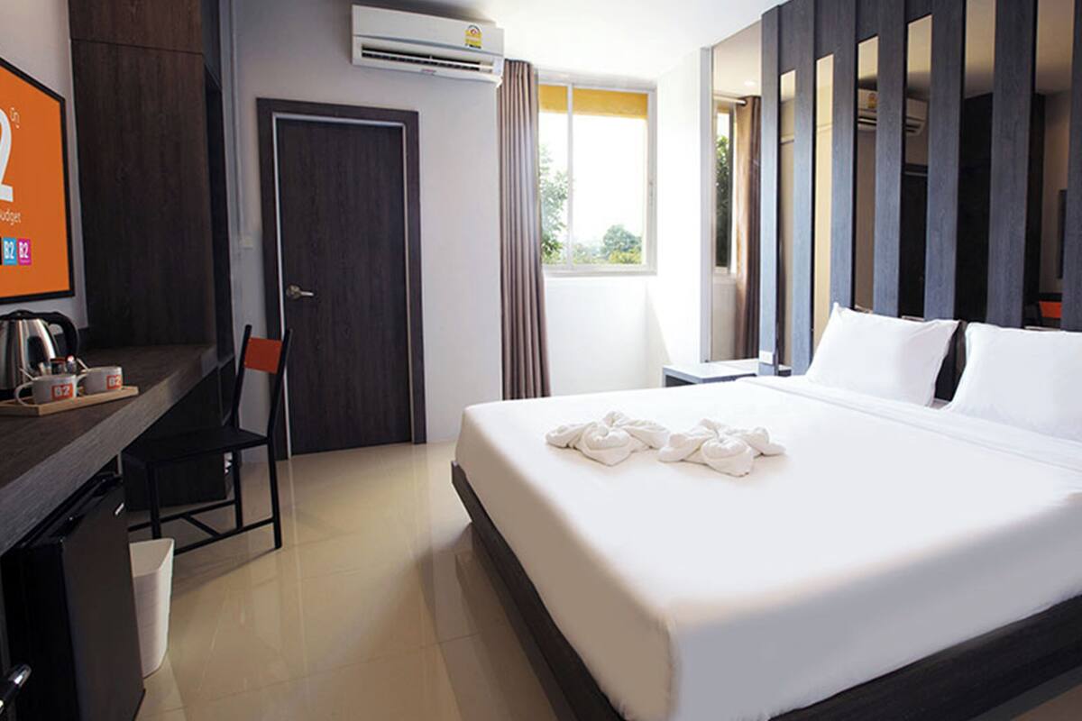 A modern hotel room with a large bed, a desk with a chair, a TV, and an air conditioning unit.