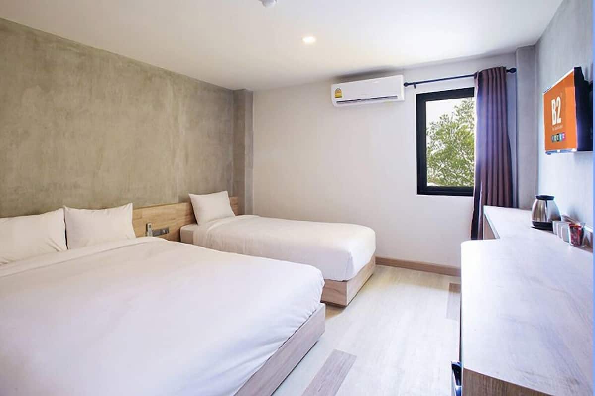 A hotel room with two beds, a window with curtains, an air conditioning unit, and a small table with a kettle.