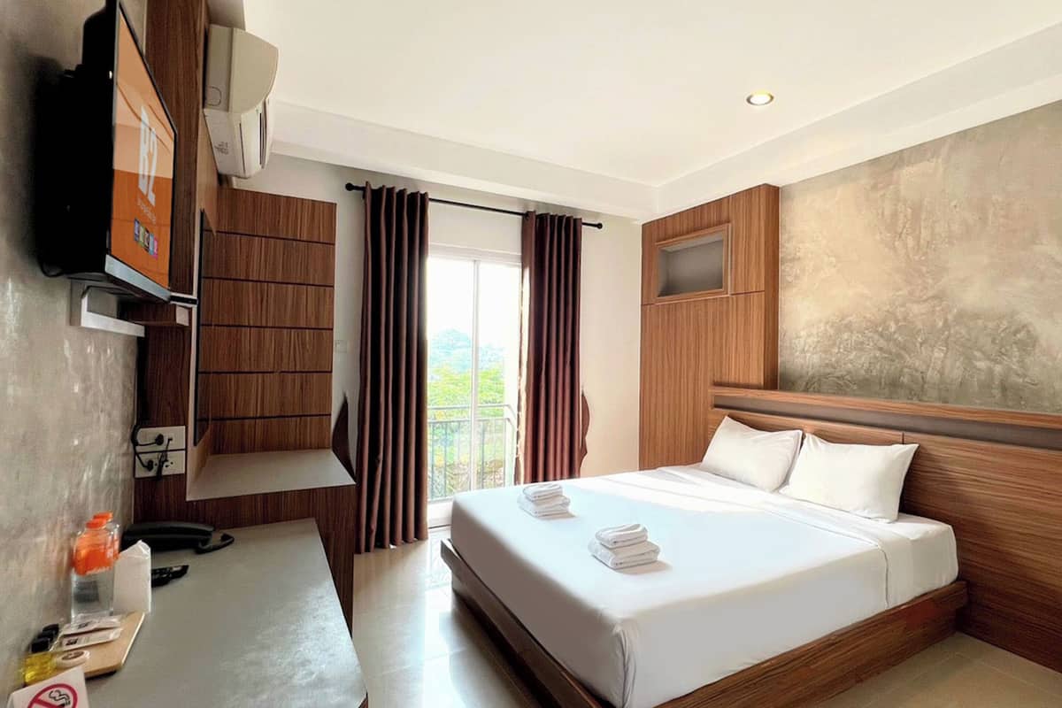A hotel room with a large bed, wooden headboard, a TV, a desk with a chair, and a window with curtains.