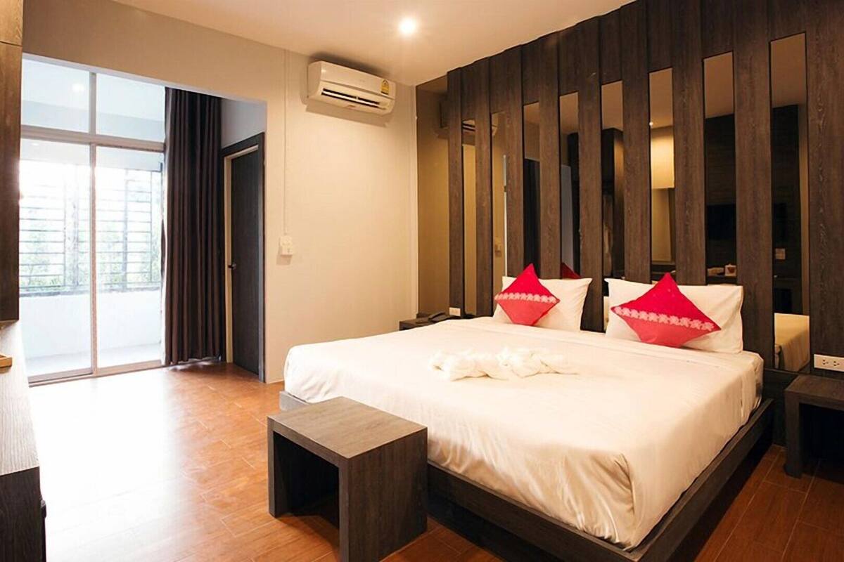 A hotel room with a large bed, a desk with a kettle, a small table, and a sliding door to a balcony.