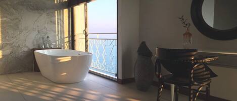 A modern bathroom with a freestanding bathtub, a large mirror, and a view of the sea through a glass door.