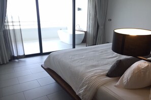Un dormitorio moderno con una cama grande, una lámpara de noche y vista a un baño a través de una puerta de cristal.