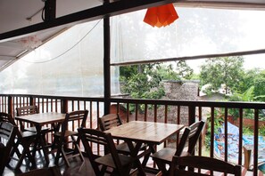 Un patio al aire libre con mesas y sillas de madera, vistas a la vegetación y un árbol, y un objeto rojo colgante.