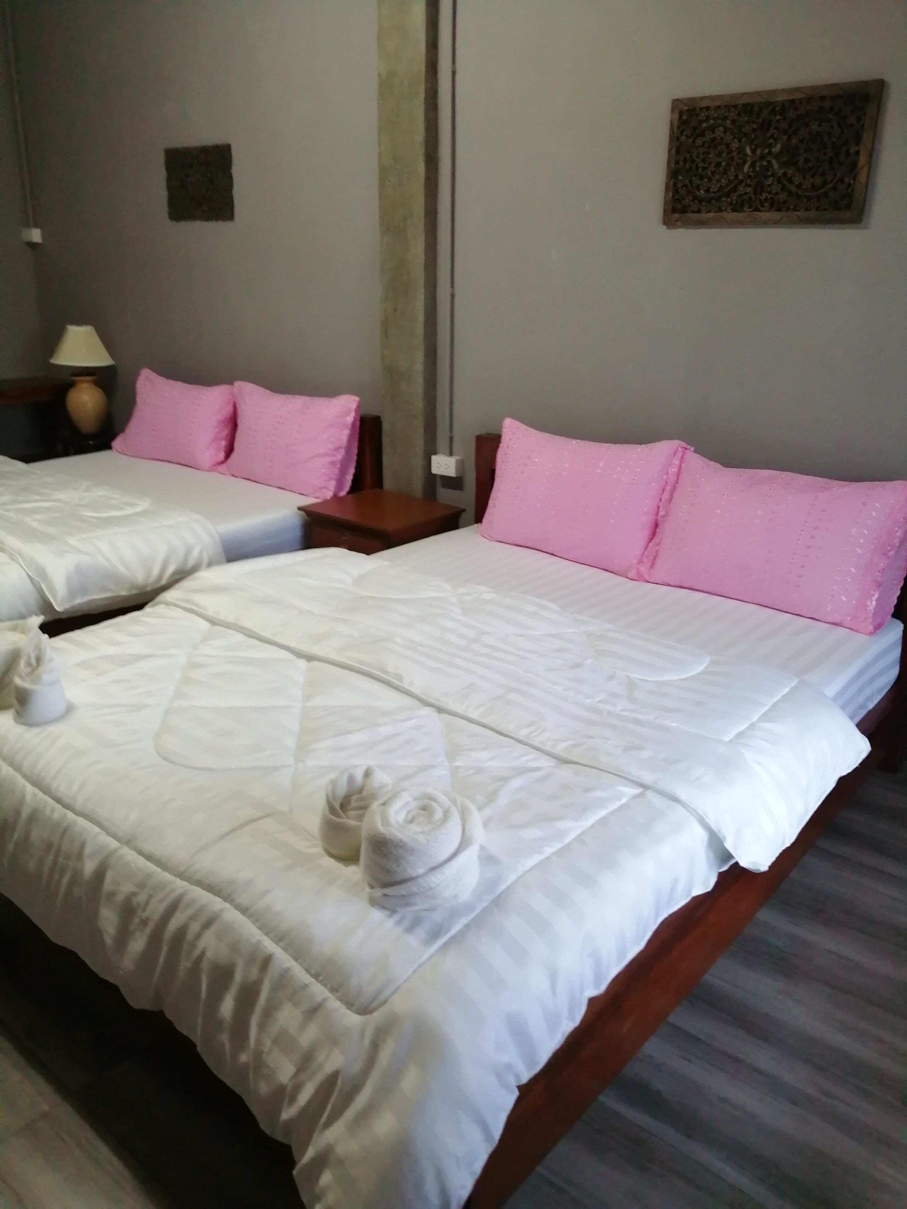Habitación de hotel con dos camas, cada una con ropa blanca y almohadas rosas. Hay una mesita con una lámpara y un adorno en la pared.