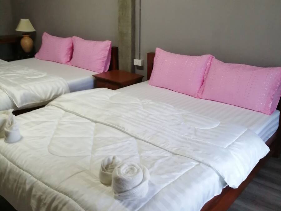 Habitación de hotel con dos camas, cada una con ropa blanca y almohadas rosas. Hay una mesita con una lámpara y un adorno en la pared.