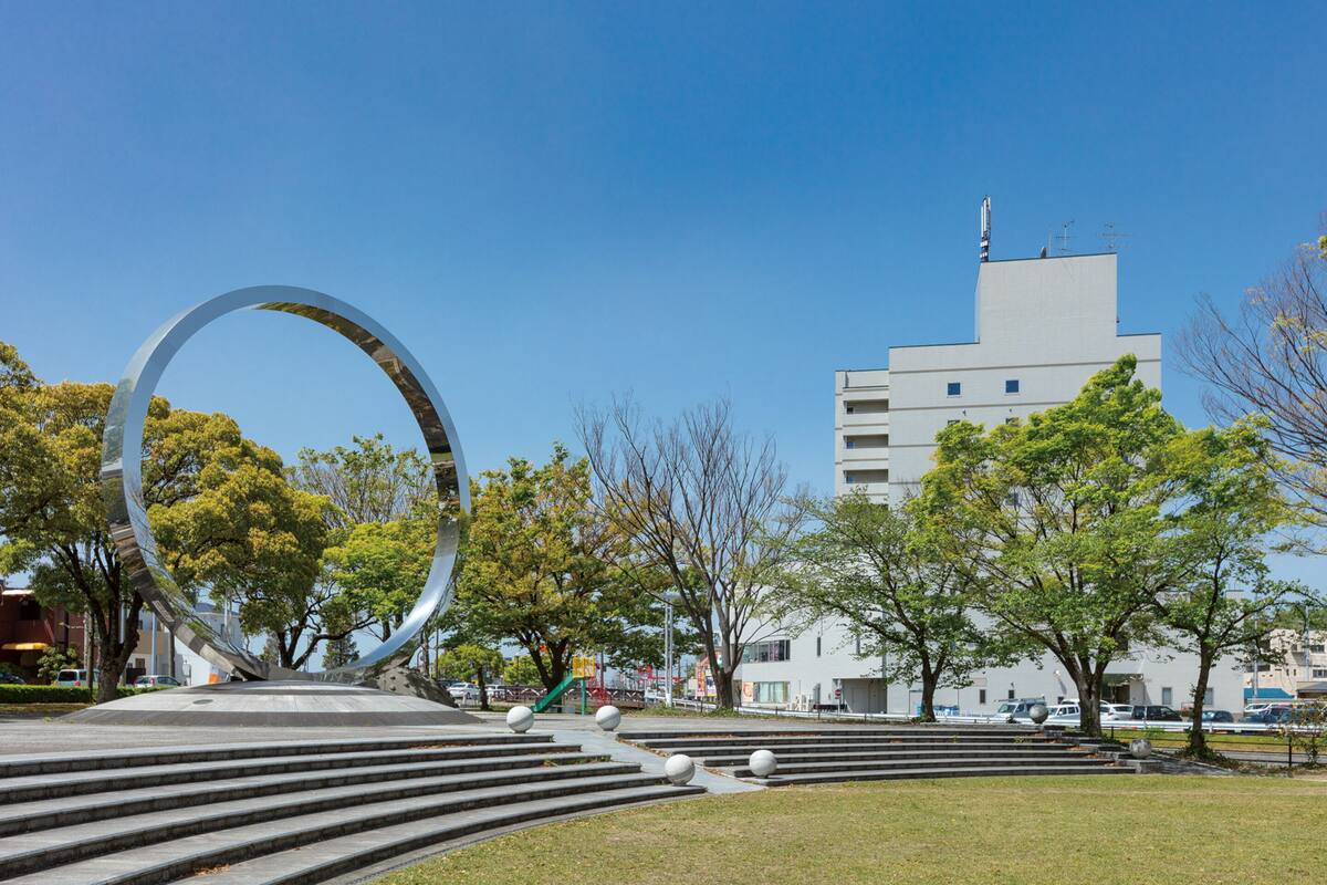 Un parc public abritant une grande sculpture circulaire, des marches menant à celle-ci et, en arrière-plan, un bâtiment moderne.
