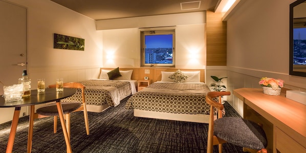 Habitación de hotel con dos camas, una mesita con sillas, un escritorio y una ventana con vista a la ciudad.