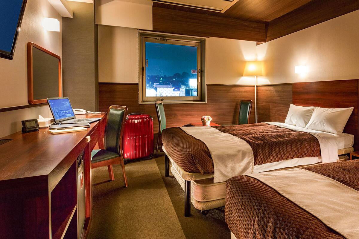 Une chambre d’hôtel avec deux lits, un bureau avec un ordinateur portable, une chaise et une fenêtre donnant sur le paysage nocturne.