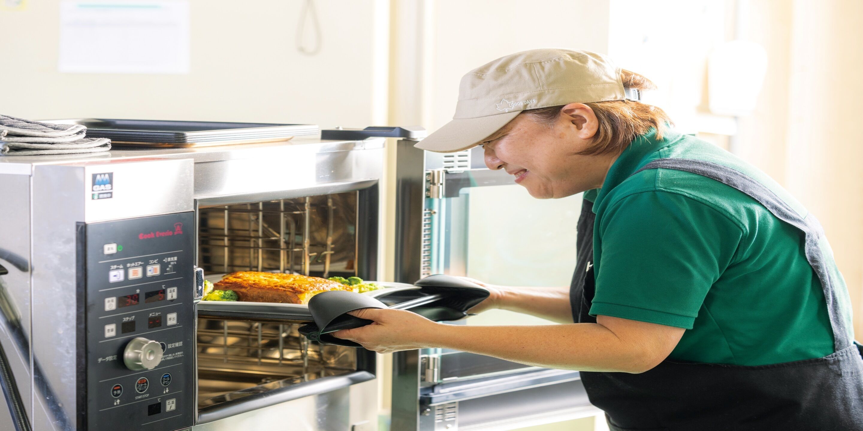 A person in a green shirt and apron using an oven mitt to handle food in a commercial oven.