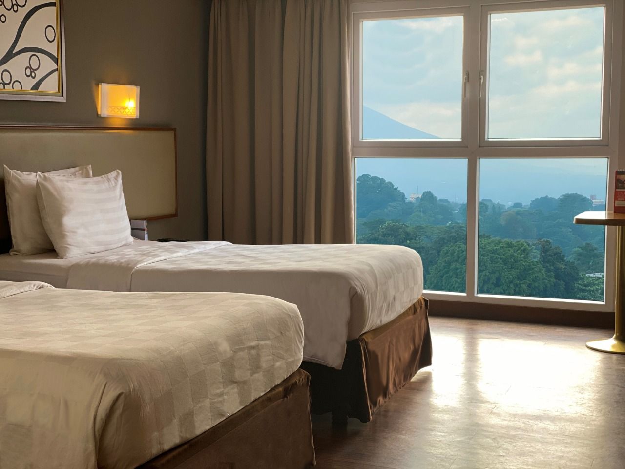 A hotel room with two beds, a large window with a view, and a bedside table with a lamp.