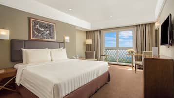 A hotel room with a large bed, a desk, a chair, a TV, and a balcony with a view.
