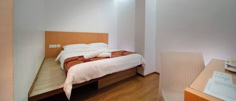 Una cama individual con cabecera de madera, colcha estampada roja y almohadas blancas. Se observa un mesita de noche de madera con una lámpara y una pequeña mesa con una taza y un platillo.