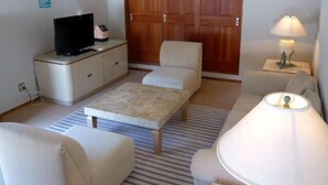 Un salon comprenant un canapé, un fauteuil, un pouf, une télévision et une porte en bois.