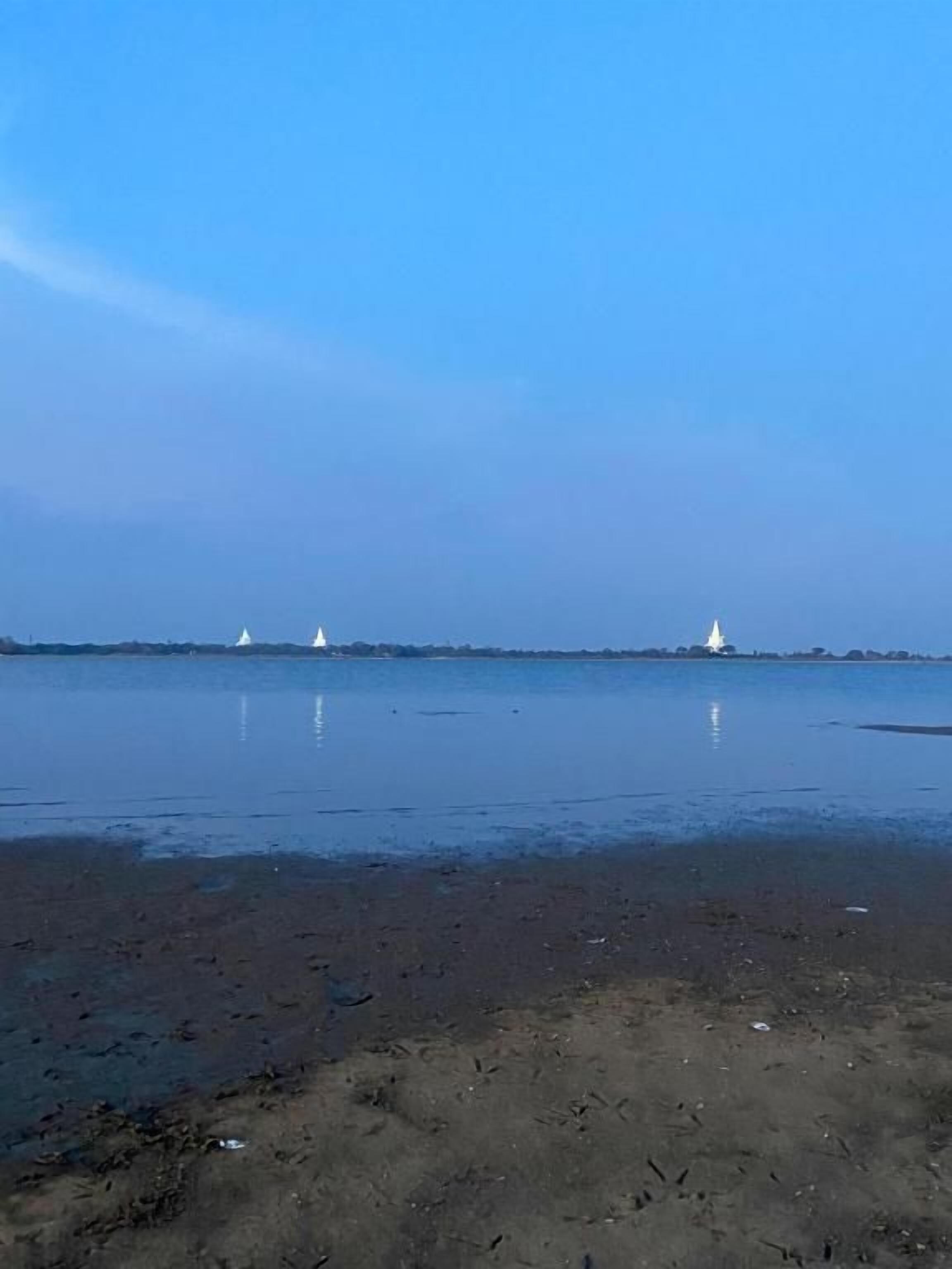 A calm body of water with a sandy shore and three white boats in the distance.