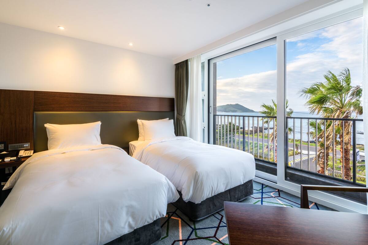 A hotel room with two beds, a balcony with a view of palm trees and a distant mountain, and a wooden headboard.