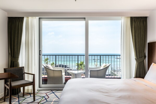 A hotel room with a large bed, a desk, and two chairs facing a balcony with a view of the ocean.