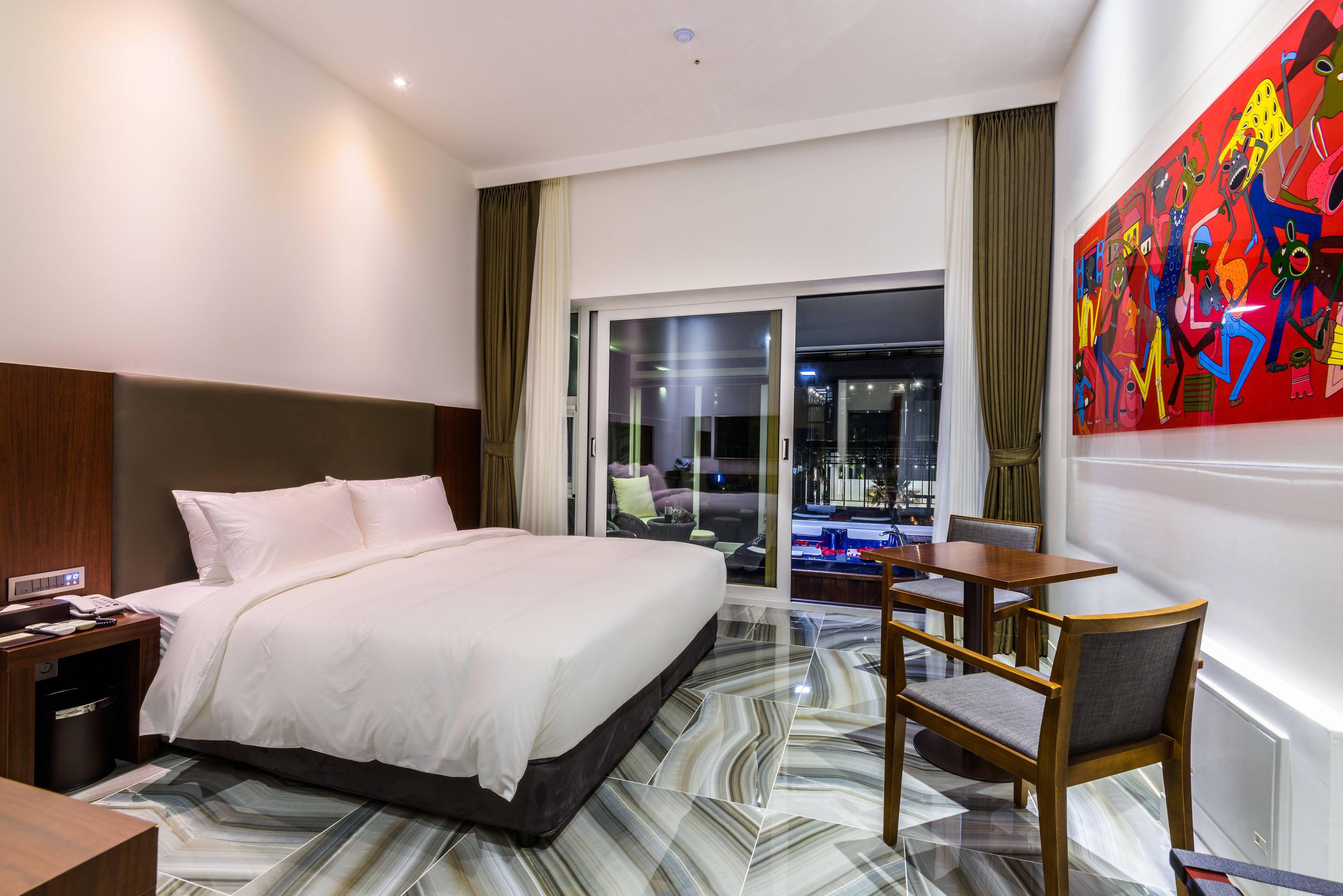 A hotel room with a large bed, a desk, a chair, and a view of the city through a glass door.