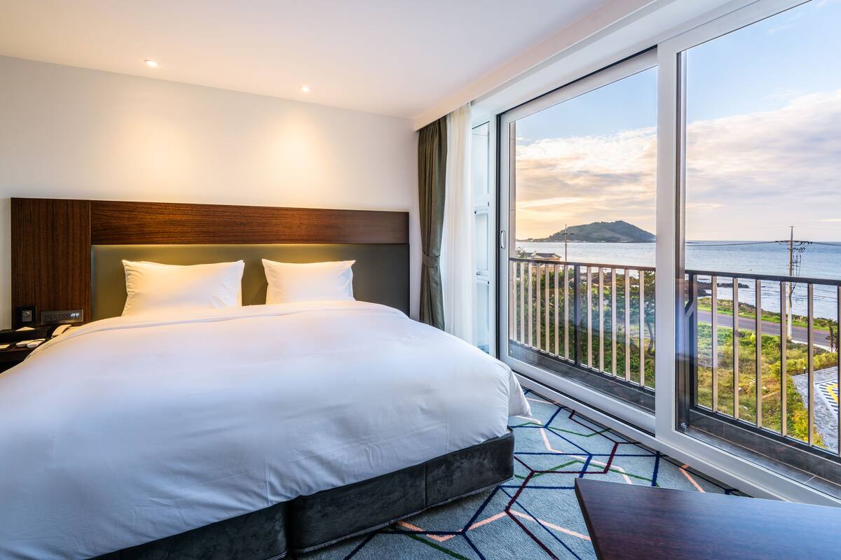 A hotel room with a large bed, a nightstand, and a view of the sea through a balcony.