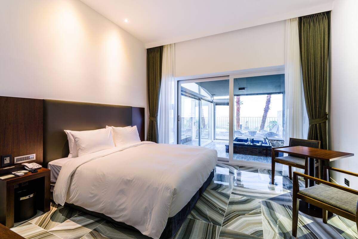 A hotel room with a large bed, a desk, and a chair. There is a window with curtains and a view of the outside.