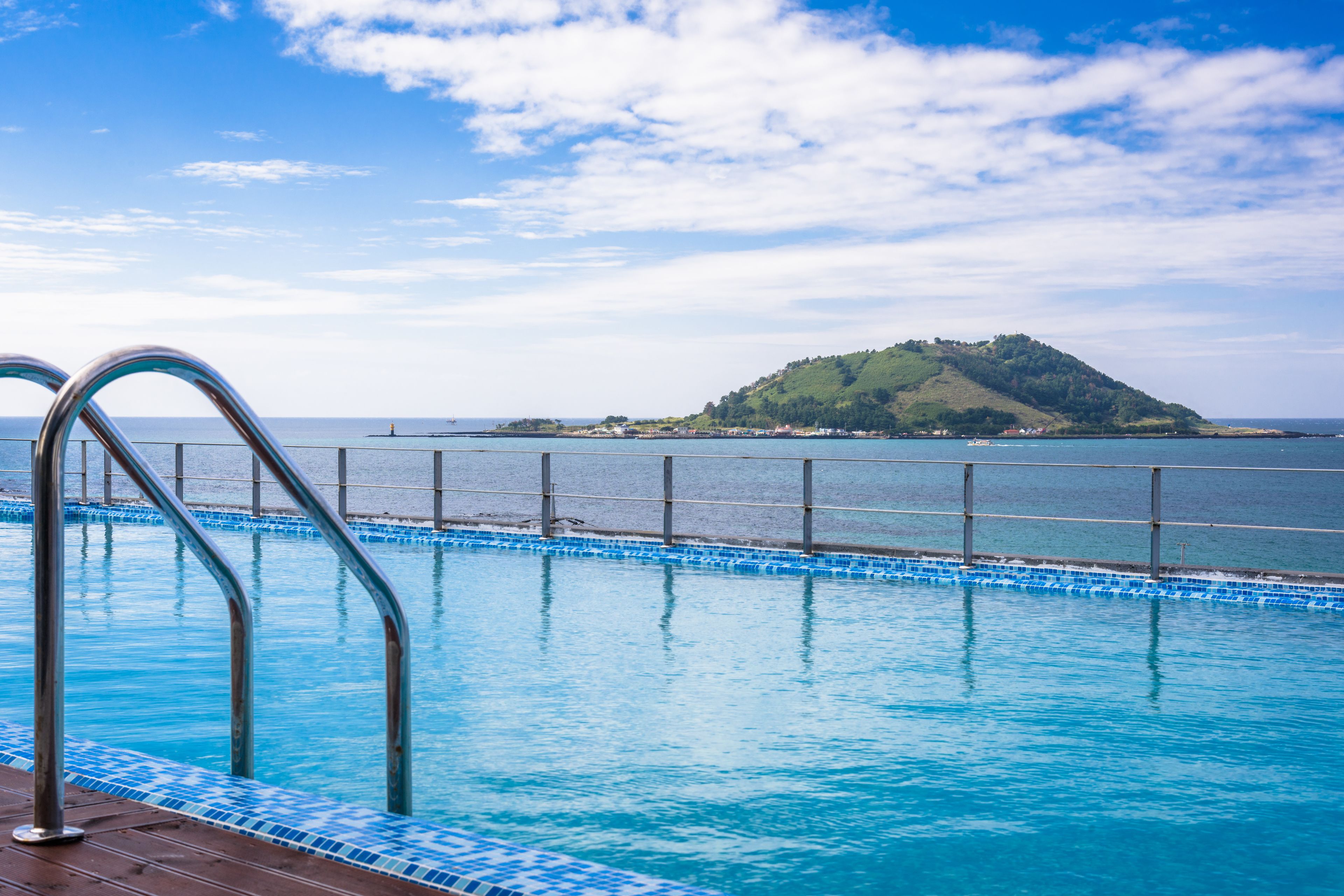 A swimming pool with metal railings, a wooden deck, and a view of a small island across the water.