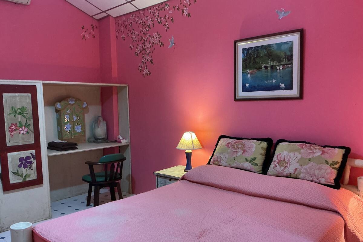 A bedroom with a bed, a desk, a chair, a lamp, and a wall with a painting.