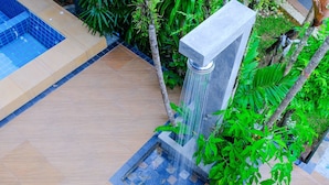 Uma área externa moderna com piso de azulejos, um pequeno espelho d'água e vegetação exuberante.
