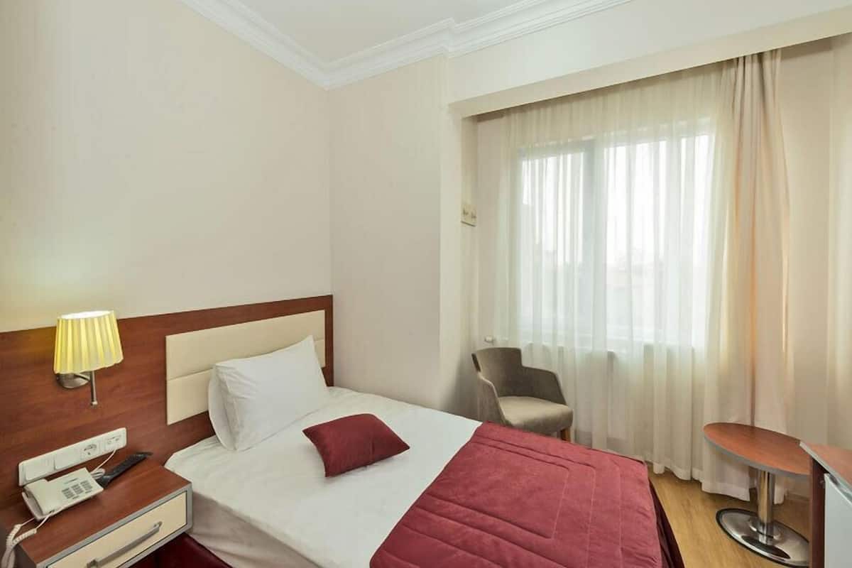 A hotel room with a bed, a bedside table with a lamp, a chair, a desk, and a window with curtains.