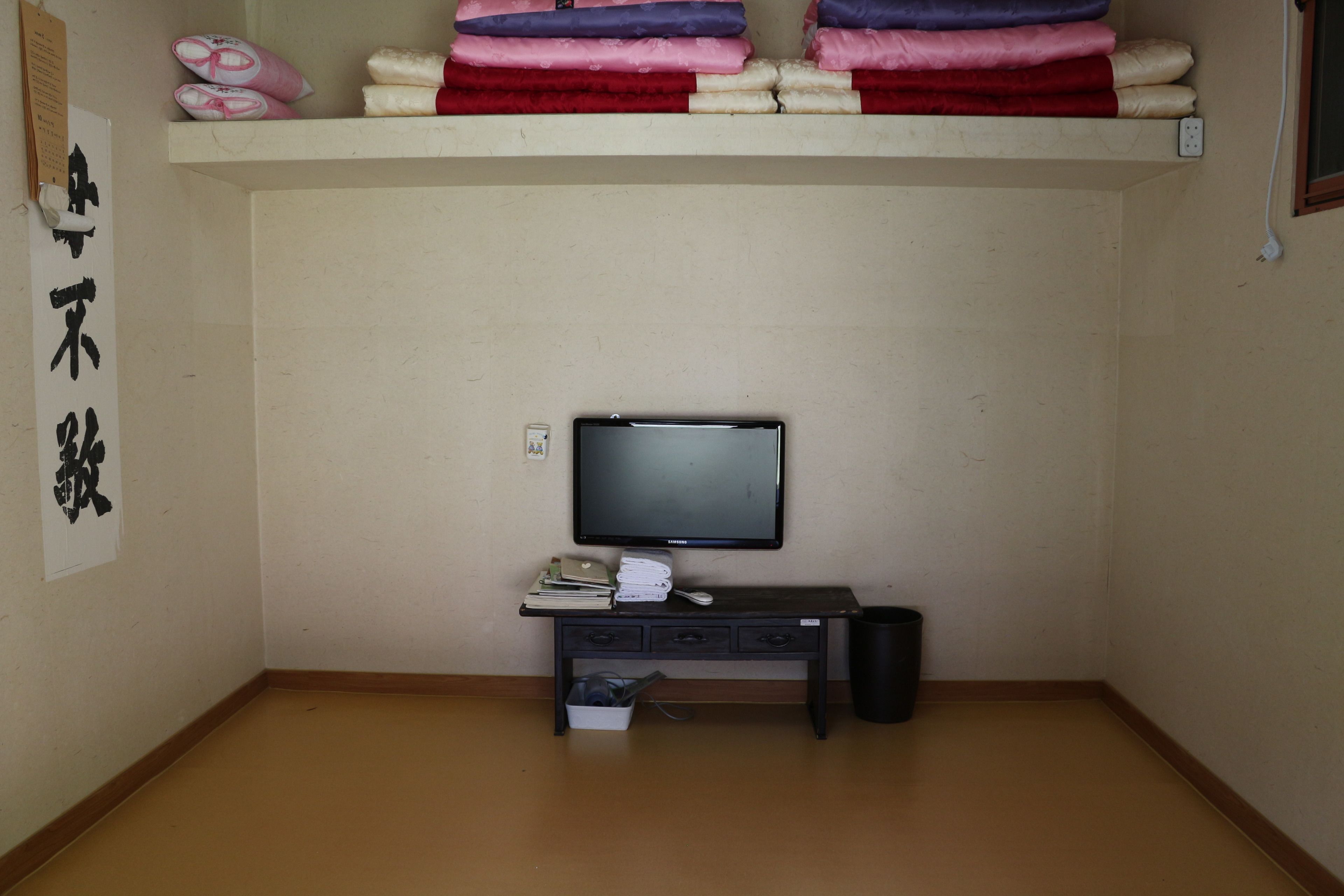 A room with a television on a stand, a small desk with books and a remote control, a trash bin, and a shelf with folded blankets.