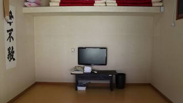 A room with a television on a stand, a small desk with books and a remote control, a trash bin, and a shelf with folded blankets.