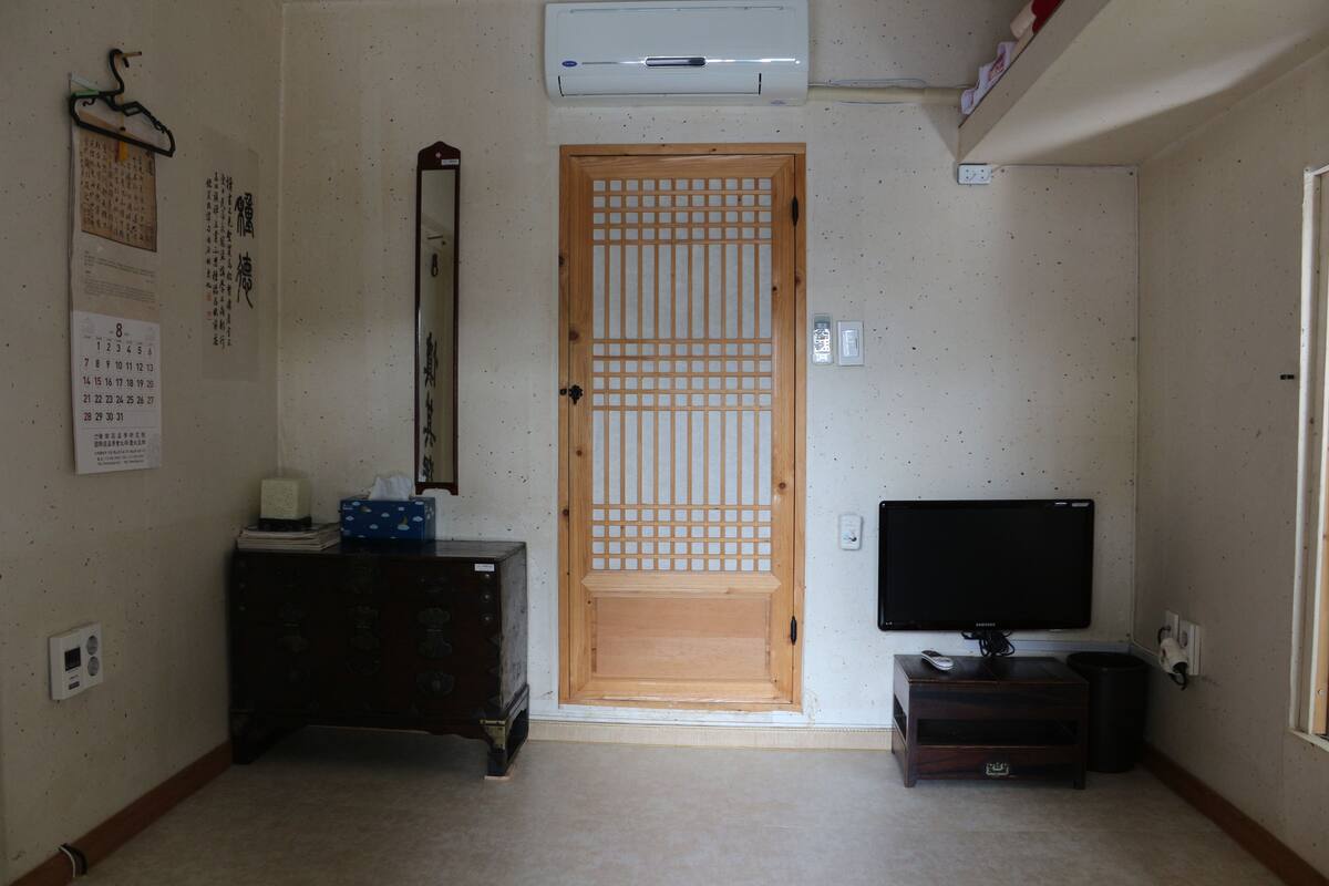 A room with a wooden door, a calendar, a small table with a box, a television on a stand, and a wall-mounted air conditioner.