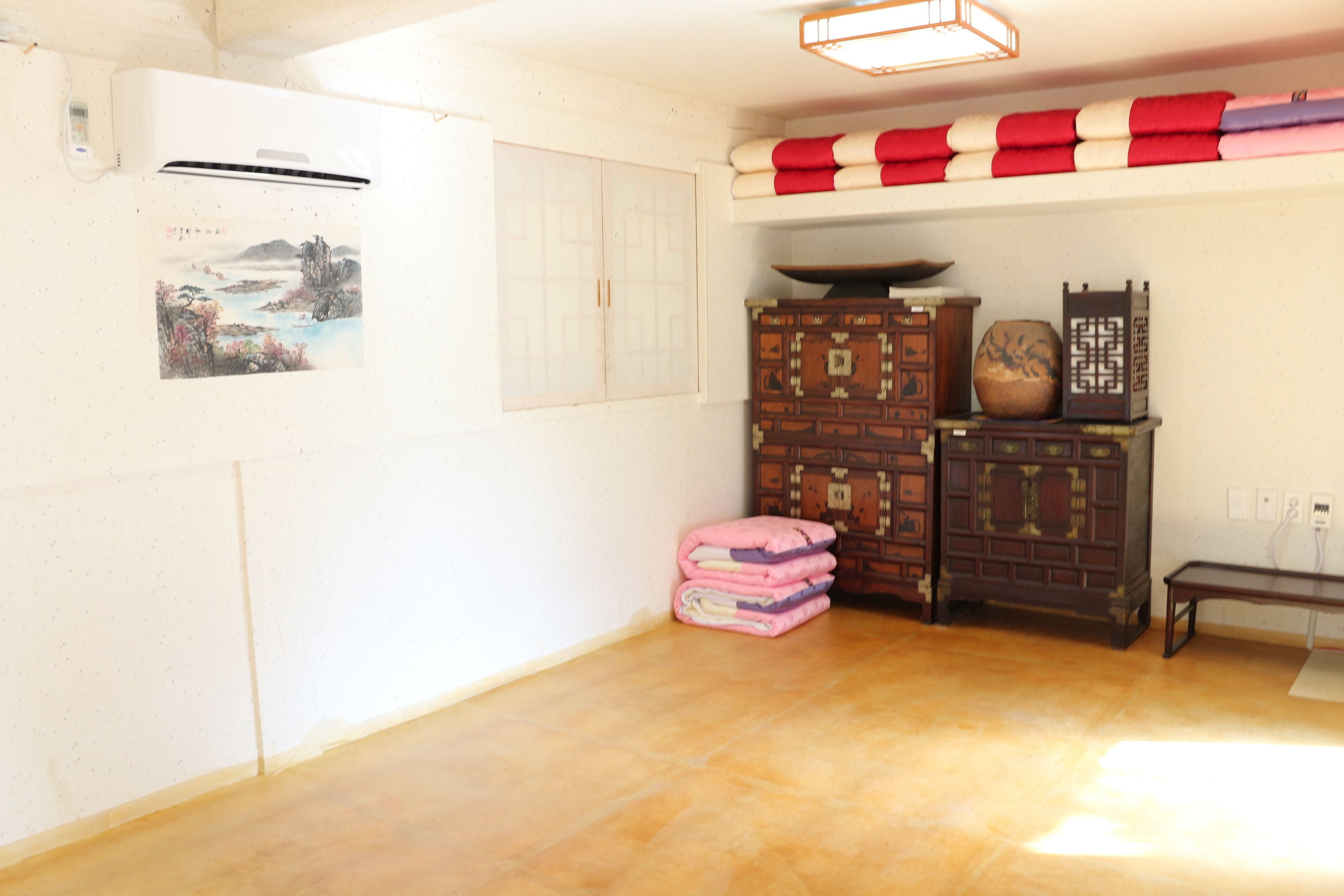 A room with a wooden floor, a wall-mounted air conditioner, traditional furniture, and a shelf with folded blankets.