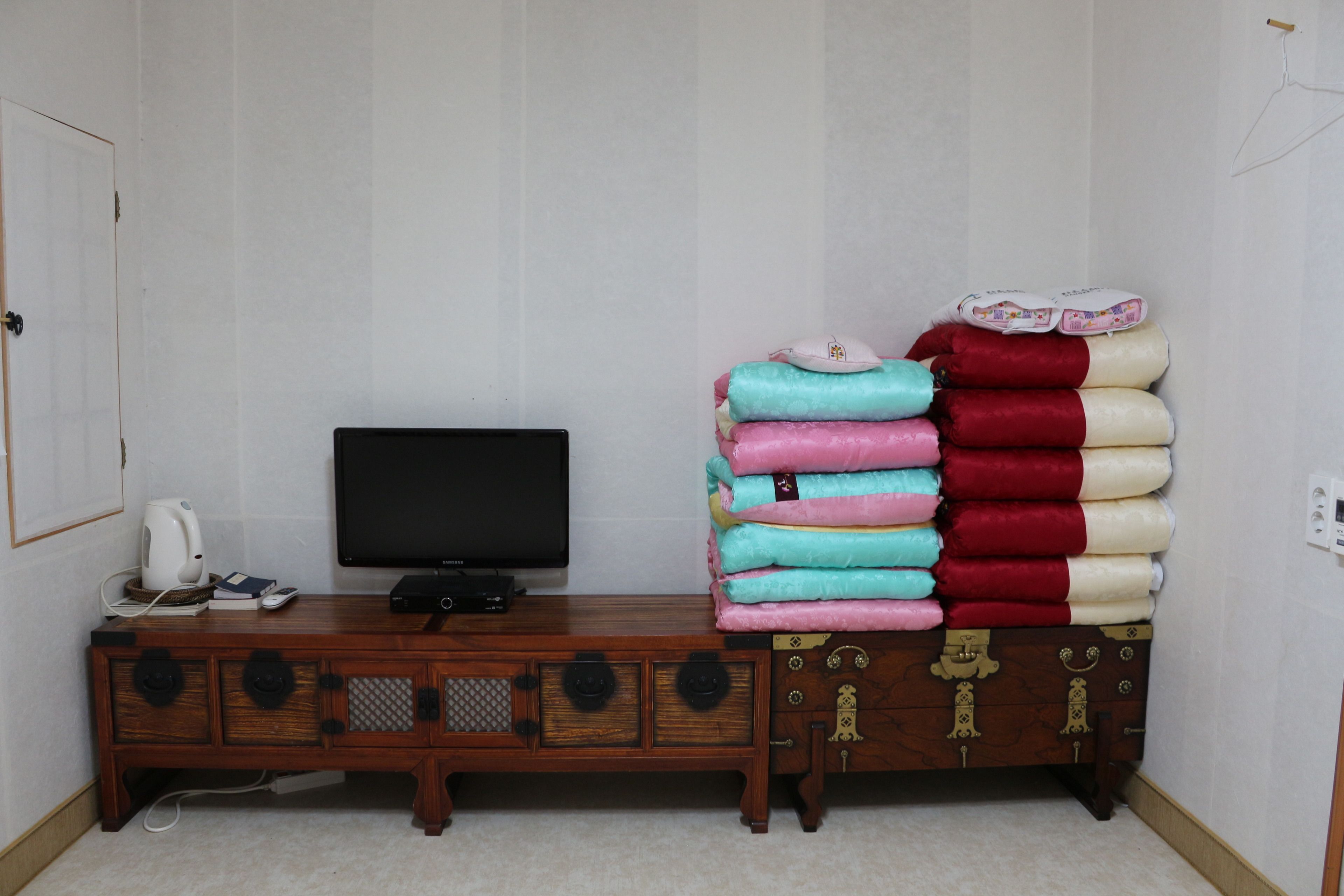 Una habitación con un televisor sobre un mueble de madera, una pila de mantas dobladas y una pared blanca.