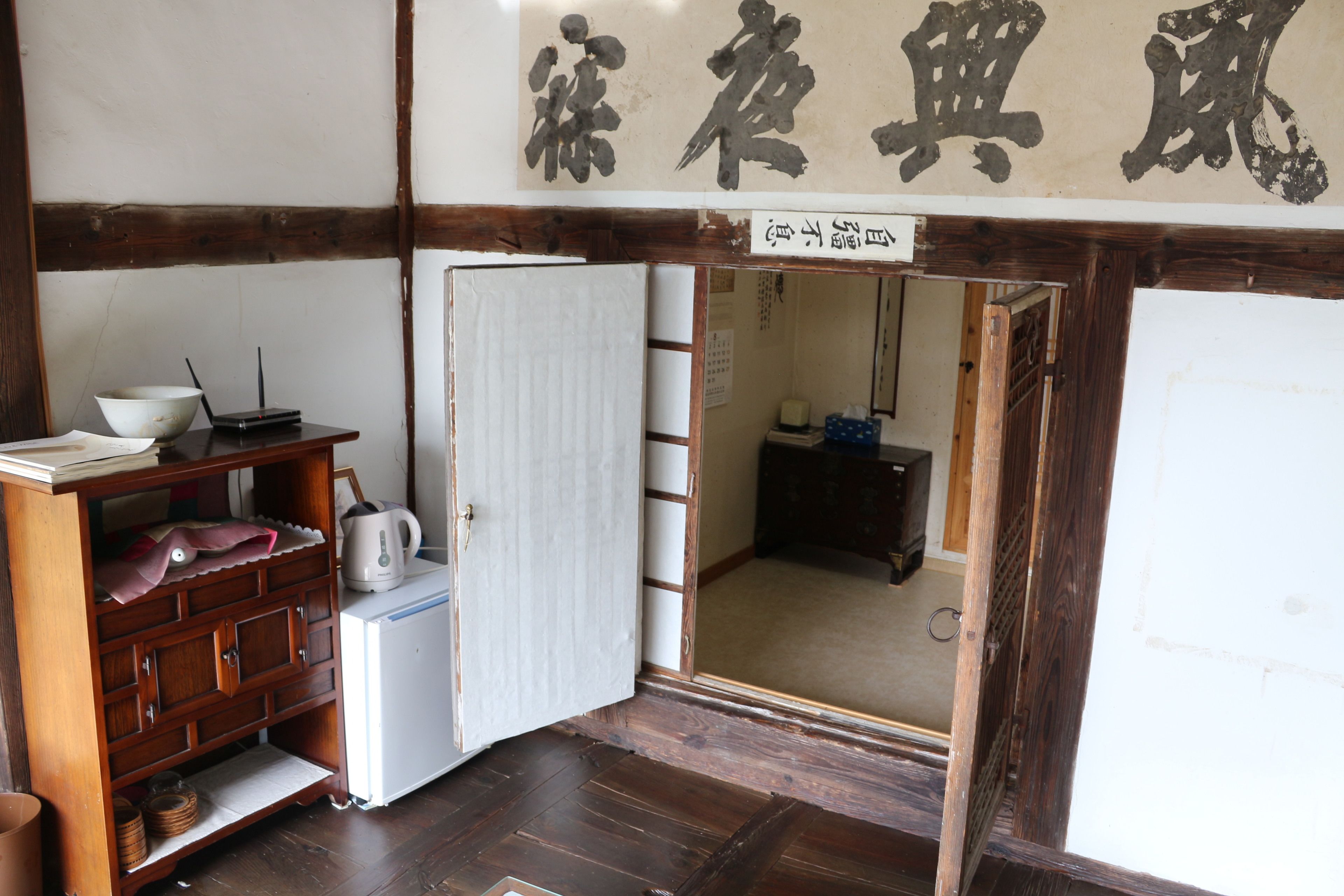 Une pièce de style japonais traditionnel, avec un sol recouvert de tatamis, un meuble en bois contenant une bouilloire, un réfrigérateur blanc et une porte coulissante donnant sur une autre pièce.
