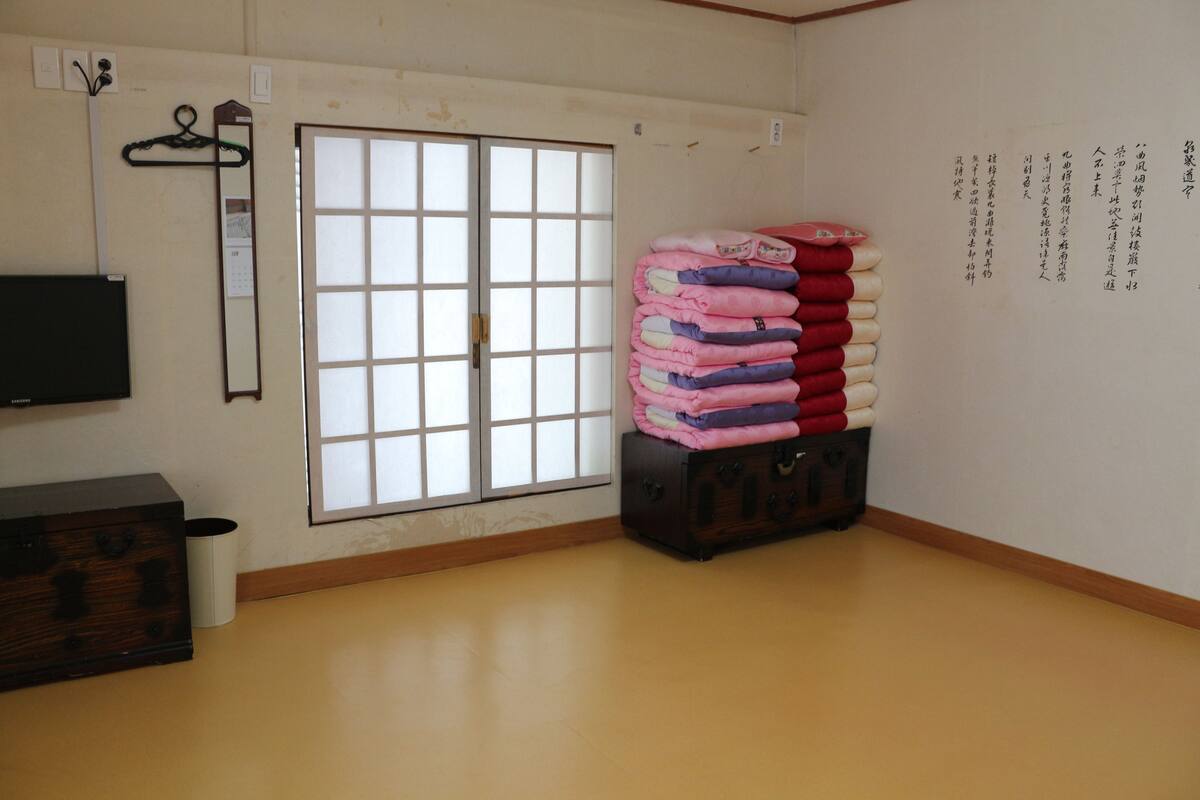 A room with a sliding door, a stack of folded blankets, a wooden chest, and a wall-mounted television.