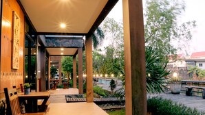 Un espacio exterior techado con bancos y mesas de madera, rodeado de vegetación y vistas a la calle.