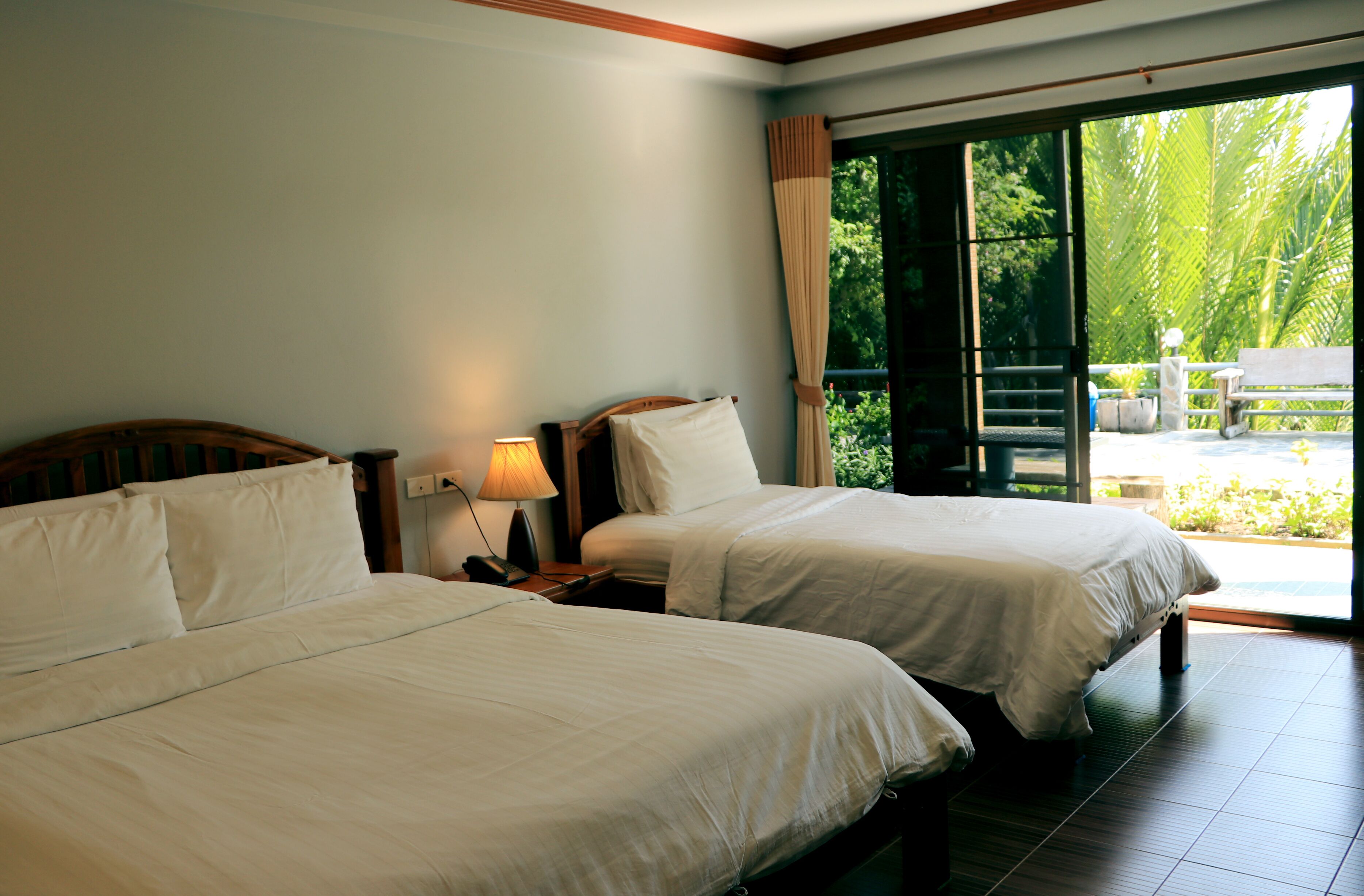A hotel room with two beds, a nightstand with a lamp, and a view of a garden through a sliding glass door.