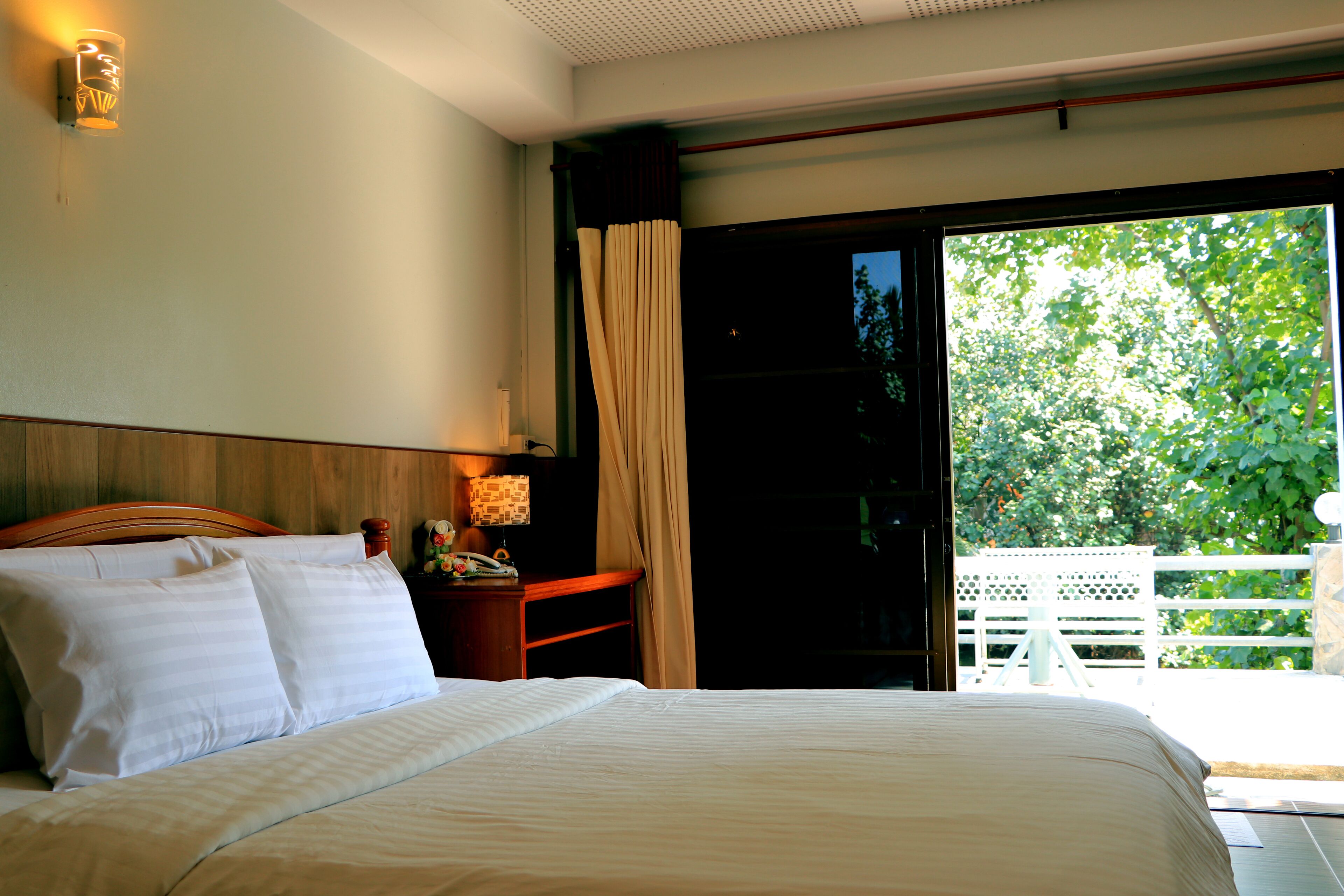 A bedroom with a bed, a nightstand with a lamp, and a view of a balcony with a white fence.