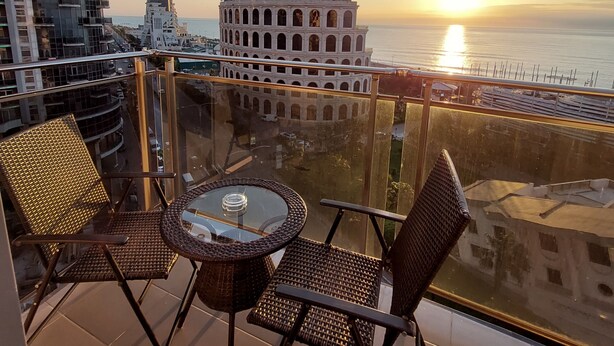 A balcony with wicker chairs and a glass table, overlooking a sunset over the sea and historic buildings. - Rock Hotel First Line (Batumi)