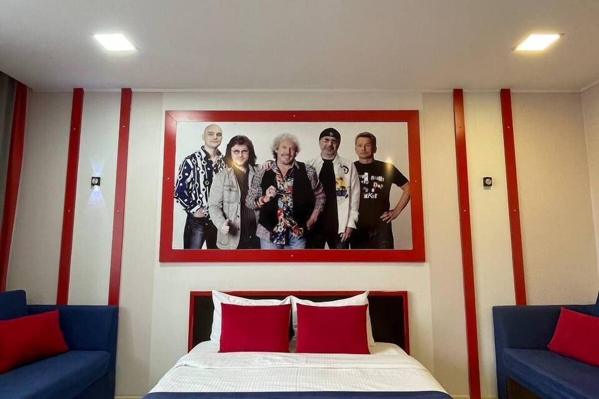 A hotel room with a large bed, two red pillows, a blue sofa, and a wall-mounted photo of five people.
