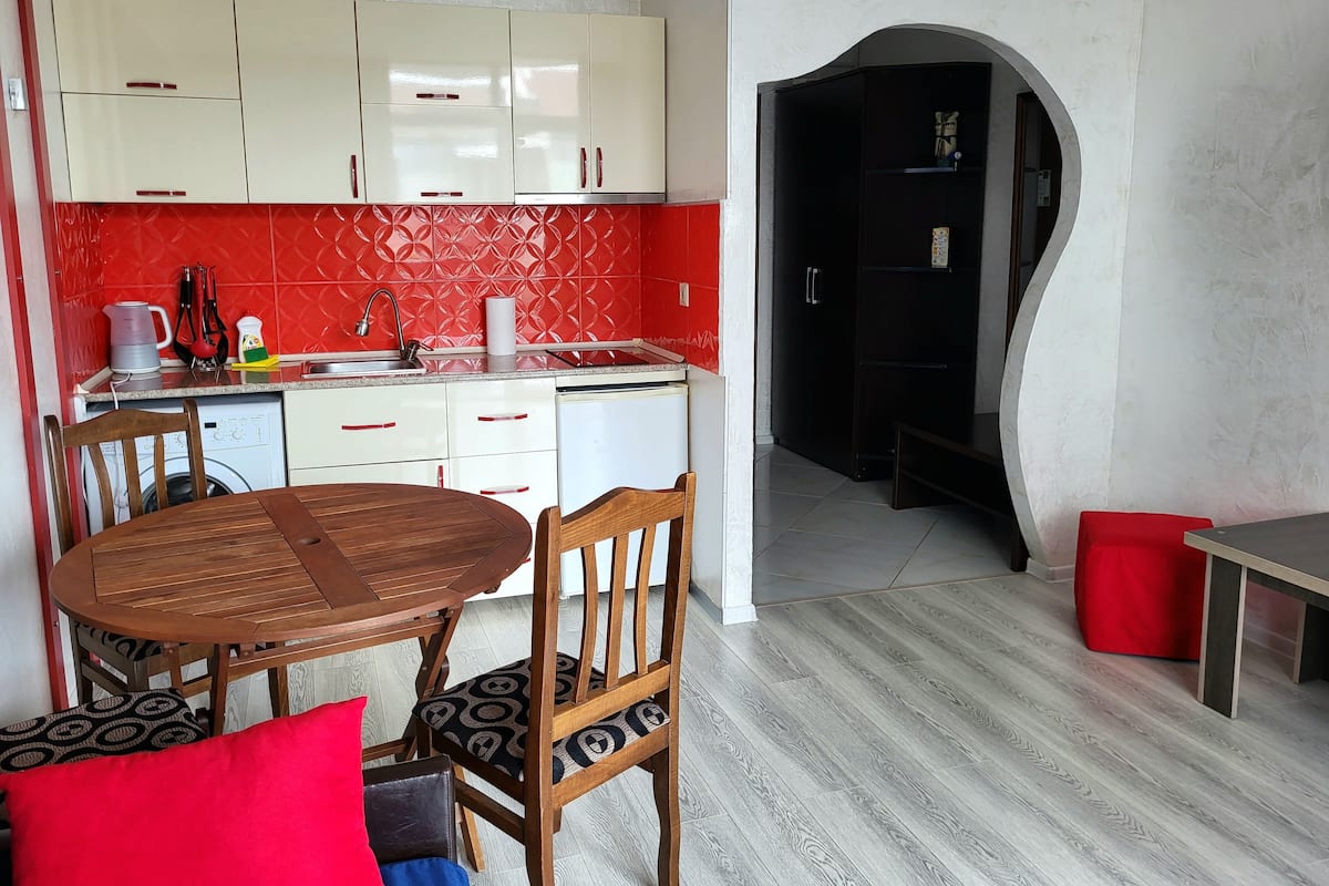 A compact kitchen with white and red cabinetry, a wooden dining table with chairs, and a red bench.