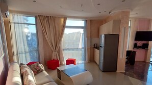 A modern living room with a sofa, a coffee table, and a kitchen area with a refrigerator and a television. - Rock Hotel First Line (Batumi)